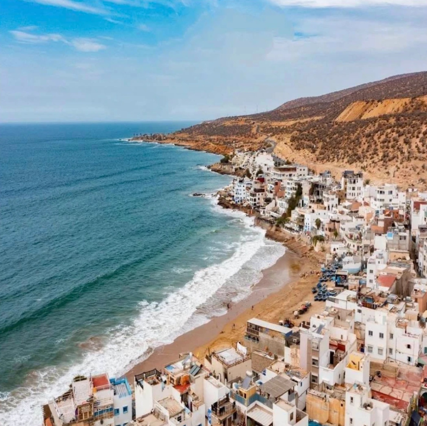 Taghazout from above: white houses along the beach, Atlantic surf rolling in, dry hills behind.