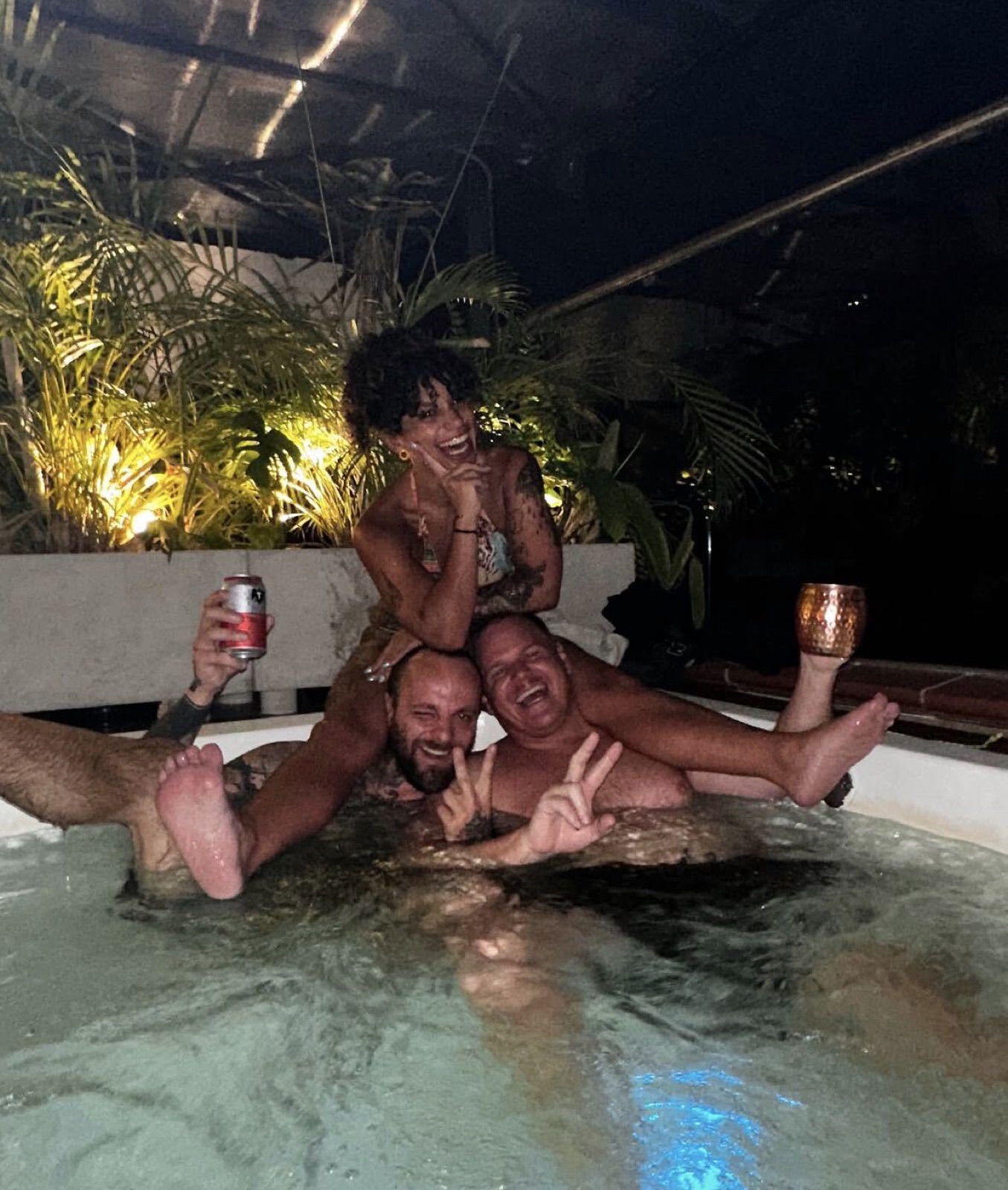 Three friends laughing in the rooftop jacuzzi at night, drinks raised.