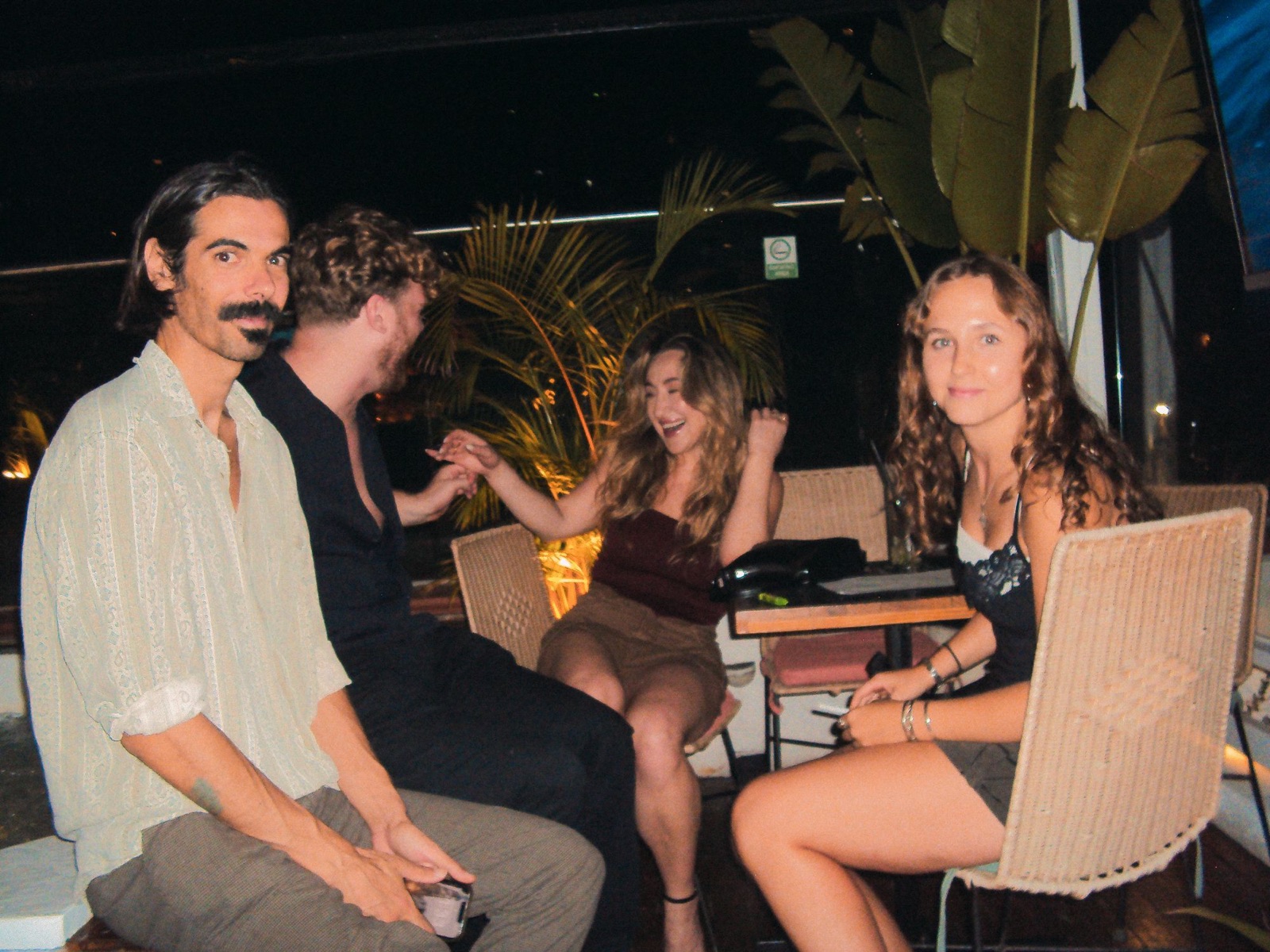 Four guests laughing together at a table on the rooftop at night, palms lit behind them.