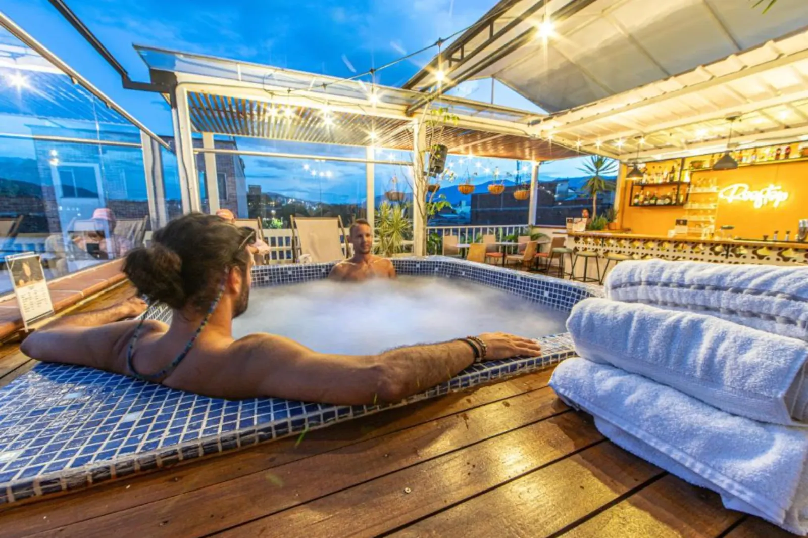 Guests in the rooftop jacuzzi at dusk, string lights on, the bar glowing behind.
